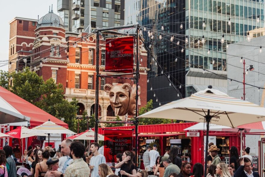 Exterior of Spiegel Haus with patrons mingling outside under umbrellas and festoon lights; city buildings in the background.