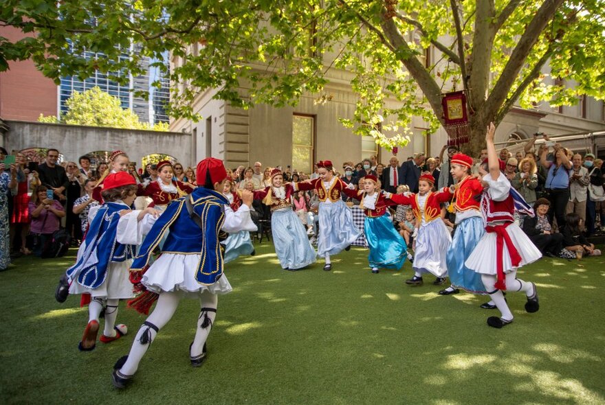 People in Greek national dress dancing in a circle in a garden with others looking on. 