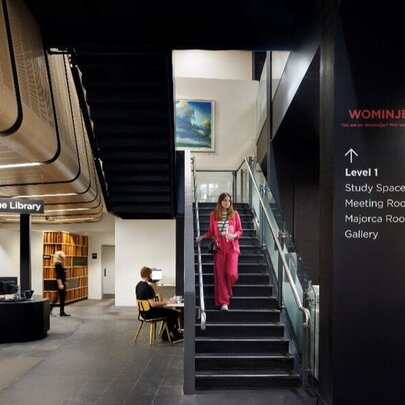 Interior view of the City Library Melbourne, featuring a modern black staircase, to the left, a bright reading area is visible with bookshelves, study desks, and people sitting at computers.