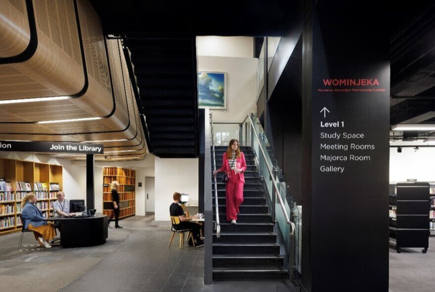 Interior view of the City Library Melbourne, featuring a modern black staircase, to the left, a bright reading area is visible with bookshelves, study desks, and people sitting at computers.