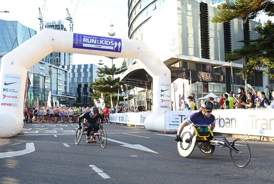 Two wheelchair cyclists cycling on a road, under the inflatable start arch of the Run for the Kids, with city buildings in the background.