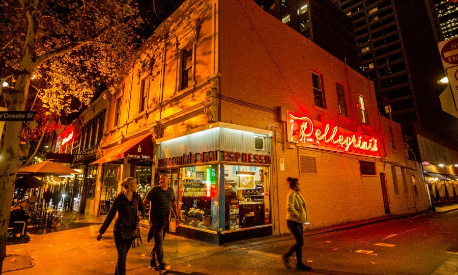 People walking past a restaurant on a corner with a glowing neon sign, at nighttime.