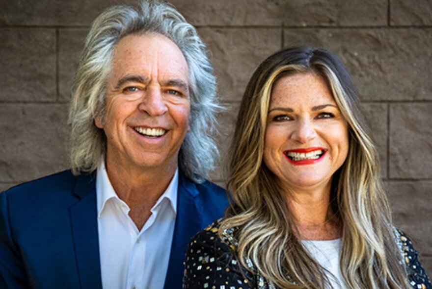 The two hosts of RockWiz, Julia Zemiro and  Brian Nankervis smiling at the viewer and posed against a stone wall.