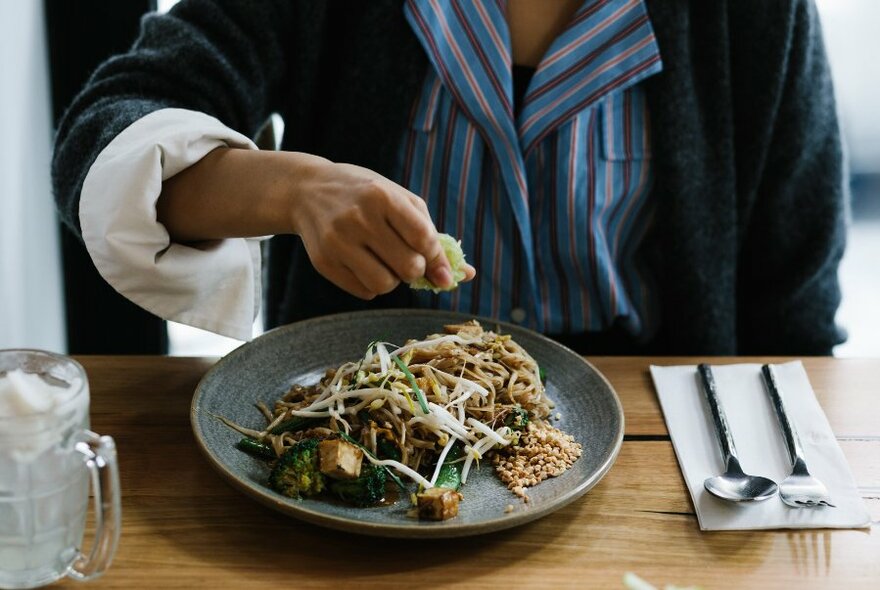 A person squeezing a wedge of lime over a plate of Thai noodles. 