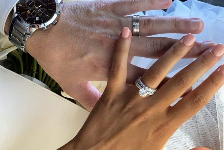 Man's hand with black-faced watch and wedding ring, beneath a woman's hand with diamond wedding and engagement rings on her ring finger.