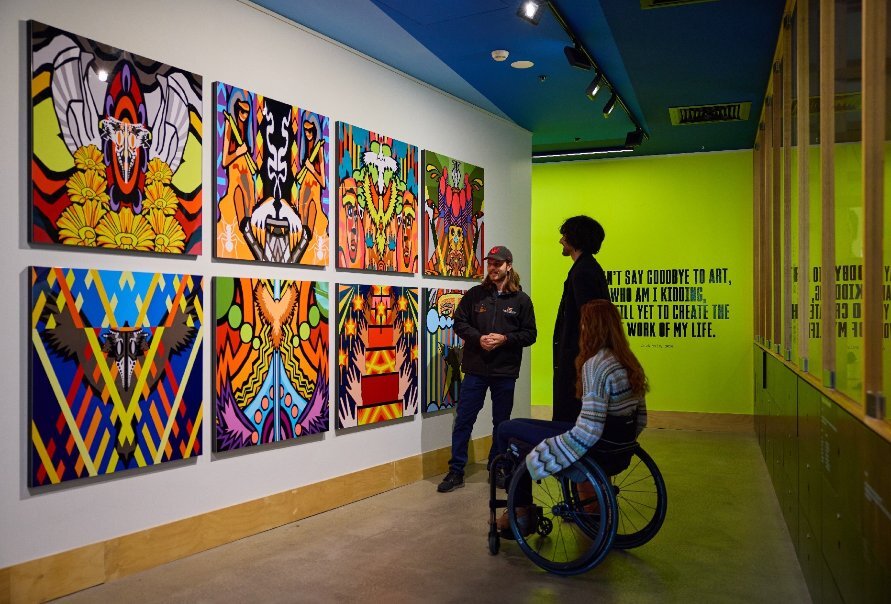 Two people, one in a wheelchair and one standing, looking at a wall of colourful art while a third person explains it to them.