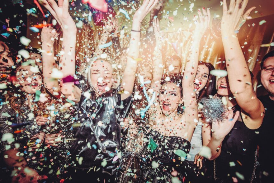 A crowd of people in a nightclub dancing and laughing with their arms up in the air and glittery confetti paper surrounding them.