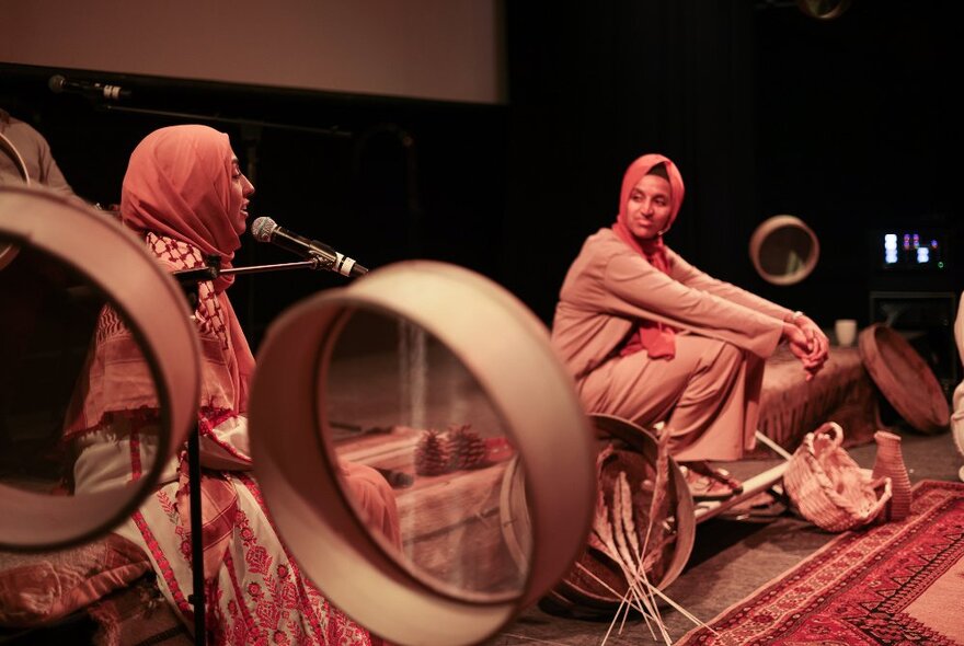 Two women wearing traditional dress seated on the floor with carpets and microphones.