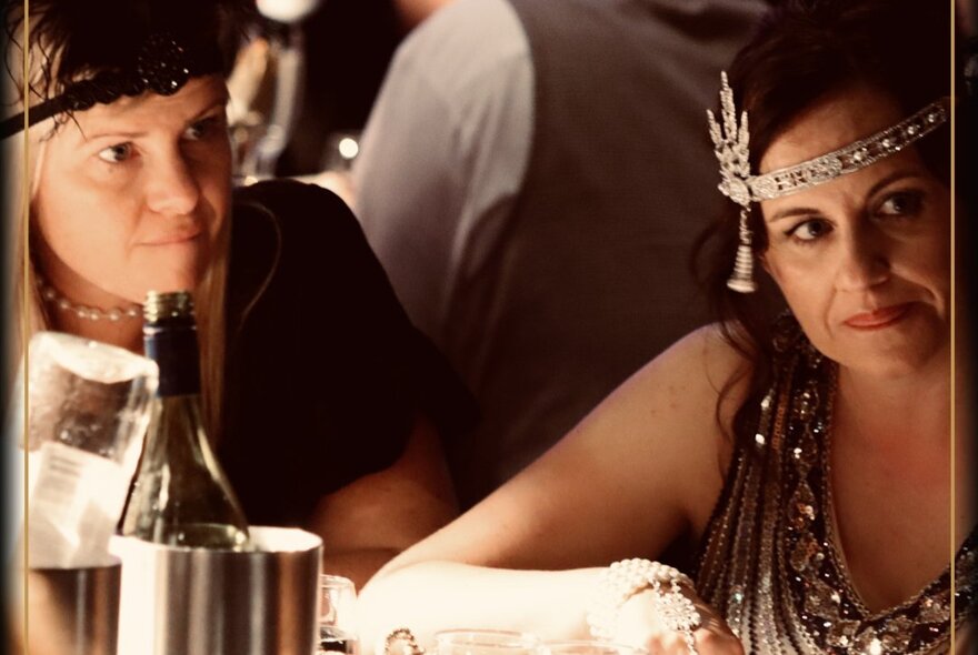 Two women dressed in Art Deco style at a murder mystery dinner, a wine bottle in a cooler on the table in front of them.