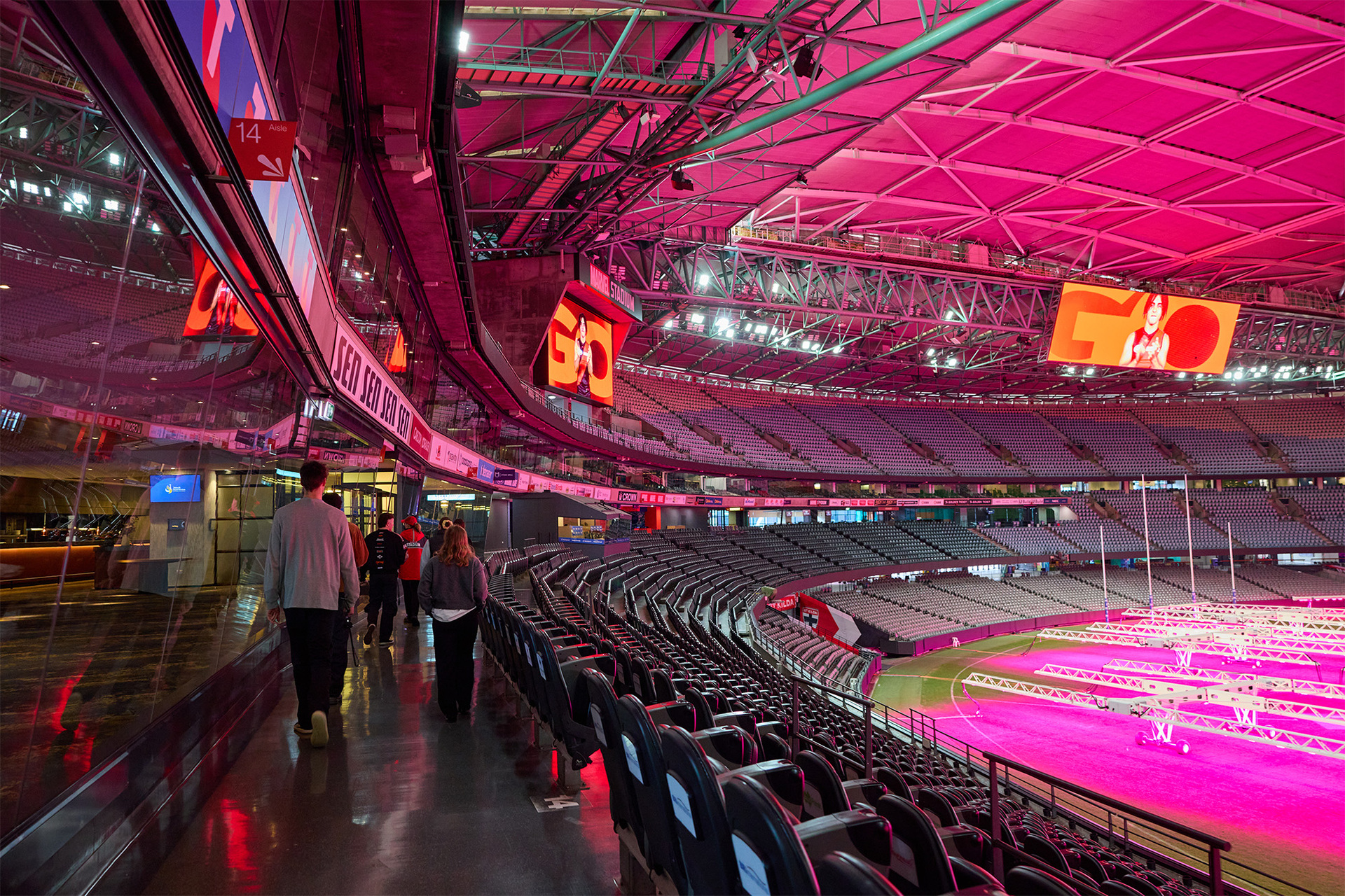 A group of people are on a tour of Marvel Stadium. They are walking past the seats in the upper levels of the stadium looking down on to the field.