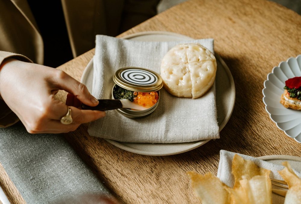 Hand scooping caviar out of a small tin on a plate with a crumpet.