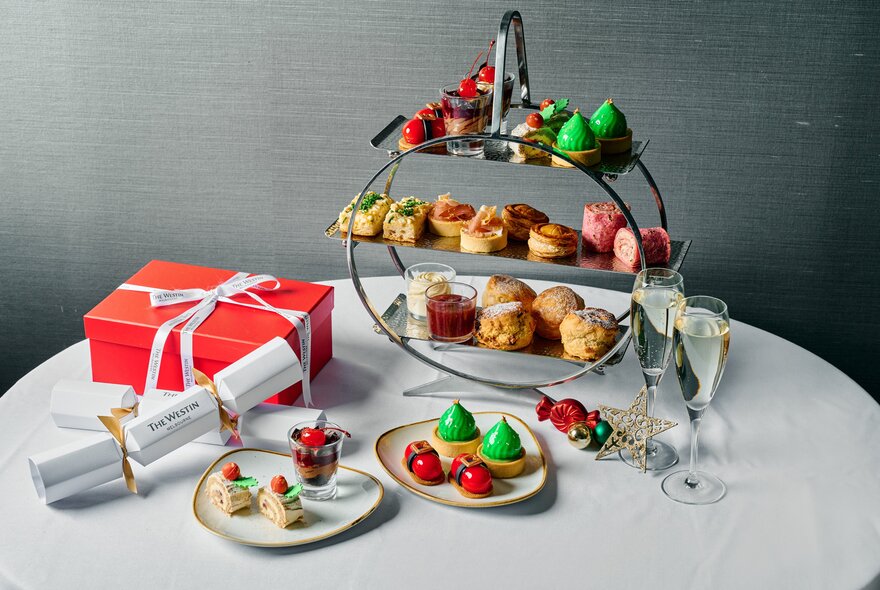 A festive-themed high tea on a round table, with a three tier stand of small items of food, a Christmas bon-bon cracker and a present in a red box.