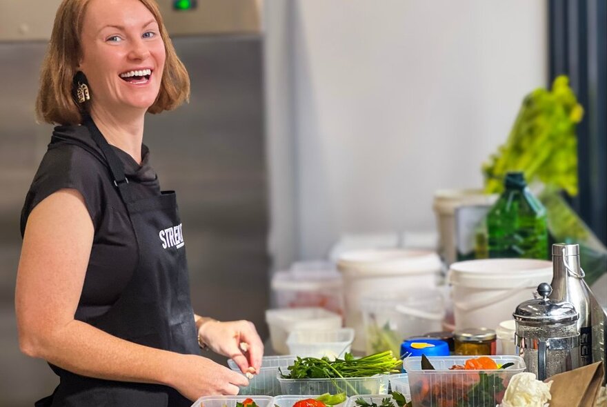 A smiling person wearing an apron with raw ingredients on the kitchen work bench in front of her.