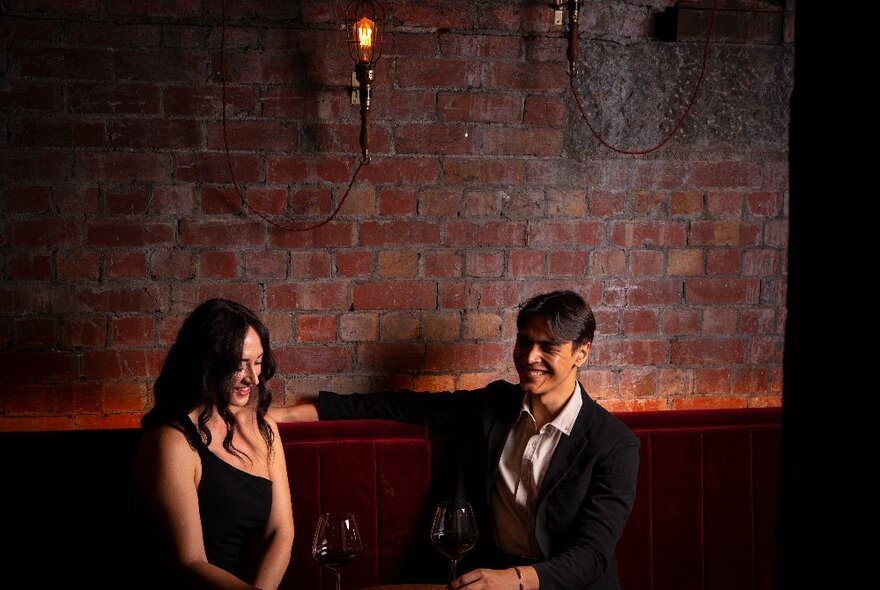 Two people smiling in a dimly-lit space with exposed brick wall. 