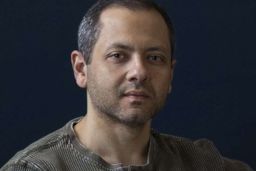 Author Omar El Akkad staring straight ahead with a neutral expression in a dimly lit area. 