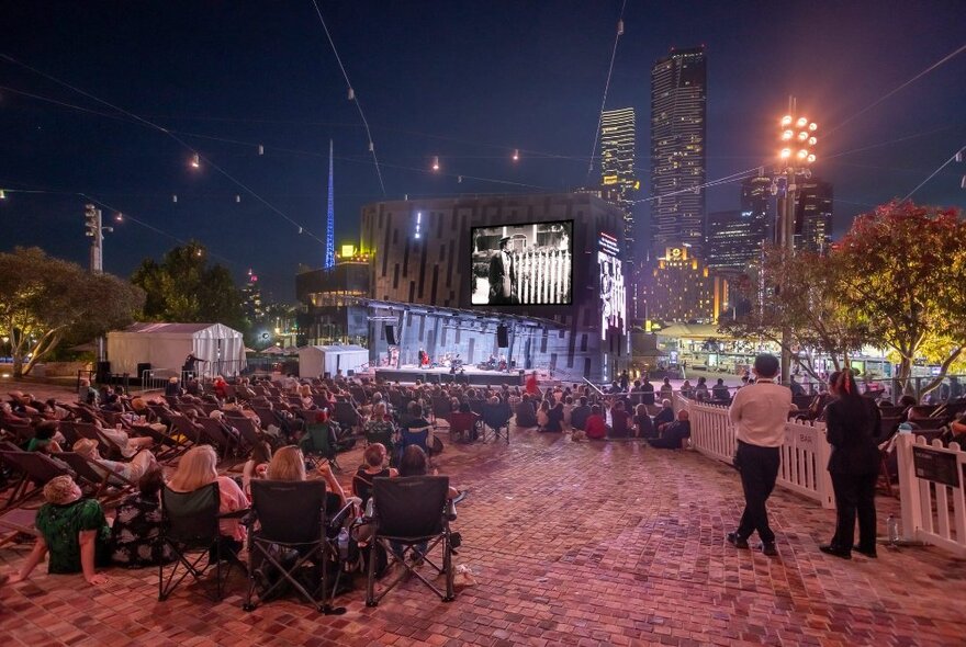 People seated on deck chairs at Fed Square watching outdoor cinema on the big screen; nighttime.