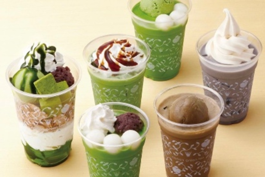 Matcha-flavoured teas and desserts, some topped with whipped cream, in plastic takeaway cups arranged on a wooden table.