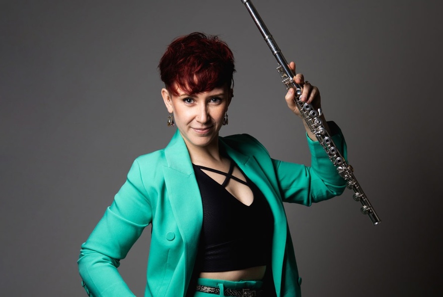 Musician with boldly cut short hair, holding a flute, wearing a turquoise jacket and strappy top.