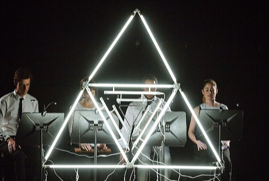 People working on computers behind a large triangular neon symbol.