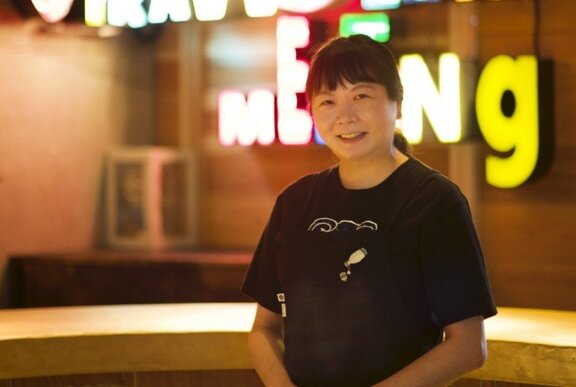 Japanese woman standing in front of neon sign, wearing a black T-shirt.