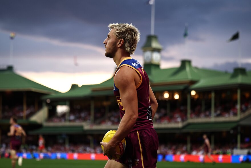 A Brisbane Lions player looks contemplative, ball in hand, at the Sydney Cricket Ground at dusk.