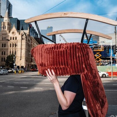 A figure walking through a Melbourne street while carrying a large, red, rocking chair over their head and shoulders, buildings and cars in the background.