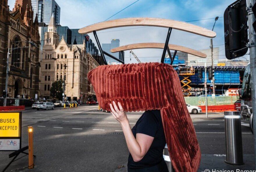 A figure walking through a Melbourne street while carrying a large, red, rocking chair over their head and shoulders, buildings and cars in the background.
