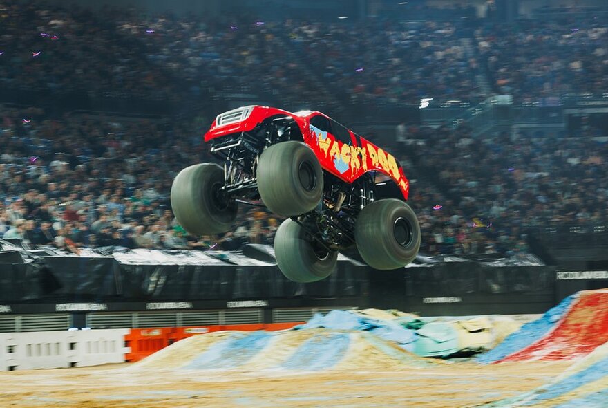 An airborne monster truck in an arena in front of a large audience.