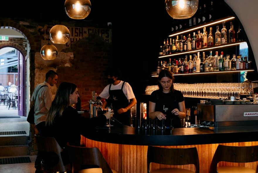 Interior of bar showing patrons sitting at a curved bar area, with bar staff and bottles of alcohol on shelves behind them.