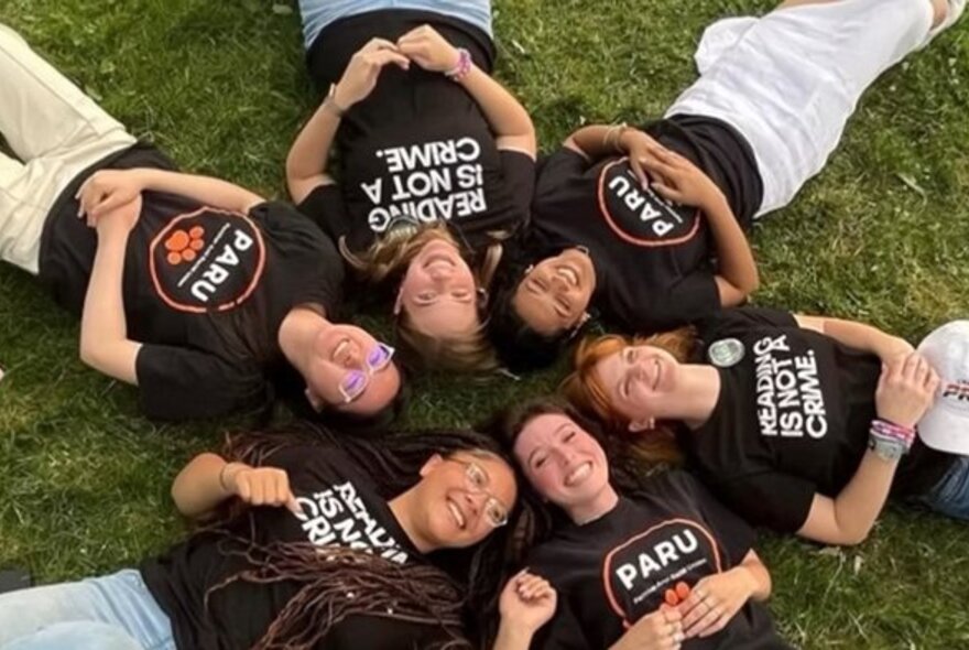Looking down at six teenagers lying on the grass on their backs in a circle with their heads touching, all wearing similar t-shirts with protest slogans on them.