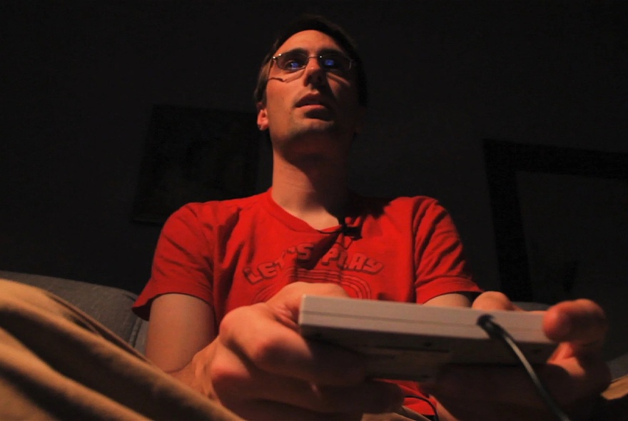 A man with glasses sits in a dimly lit room, wearing a red t-shirt holding a game controller with both hands.