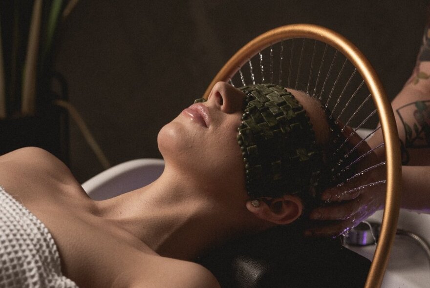 A woman in a spa, lying on her back with a woven seaweed mask over her eyes and a bronze ring providing a water massage for her head.