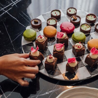 A high-angle shot shows a hand reaching for a petit four on a checkered platter filled with various colourful sweets. 