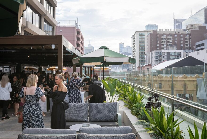 The rooftop bar at Bossley with couches, plants, shade umbrellas and people socialising with drinks; views of the city in the background.