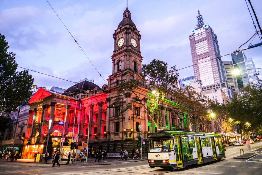 A tram going past a city building with a clock tower, with pink and green lights on the building.