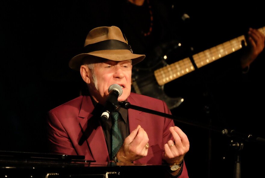 Musician, Stewart D'Arrietta, singing into a microphone and clicking his fingers while seated at a piano.