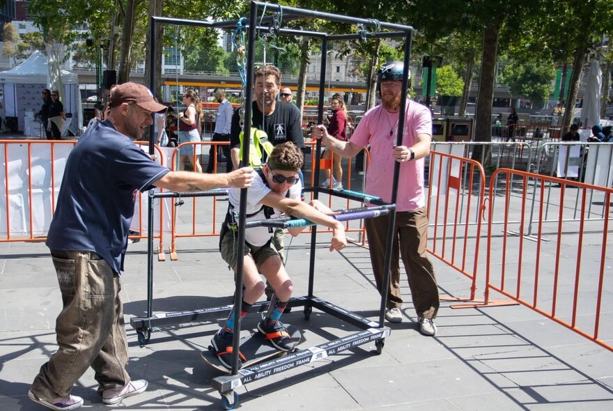 A person using an adaptive skate frame with the support of supervisors to experience skateboarding at a skate park. 