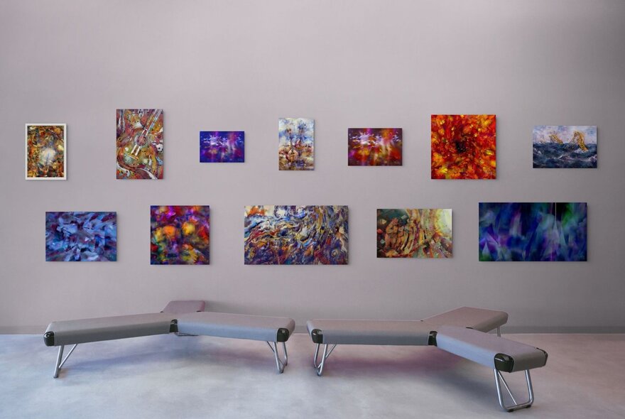 A gallery space with many colourful smaller paintings on the wall and two tri-shaped seats close to each other. 
