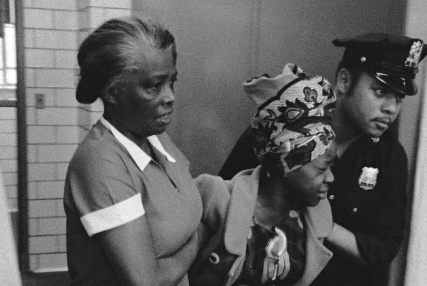 A black and white image of a woman behind held up by another woman on one side and a policeman on the other. 