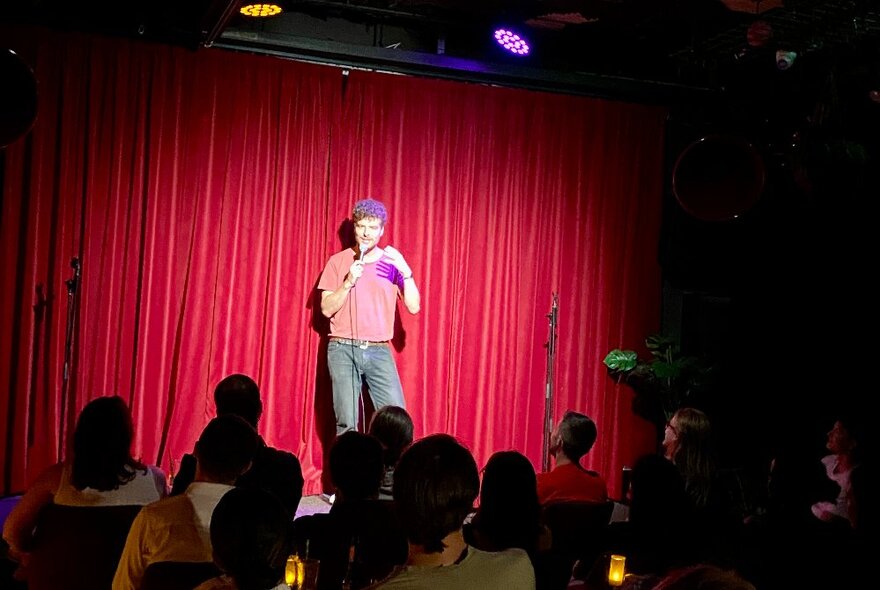 Comedian standing with a microphone on stage in front of red curtains and seated audience.