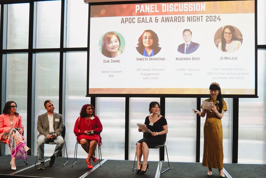 A panel of speakers seated on stage underneath a projector screen showing their faces and names.