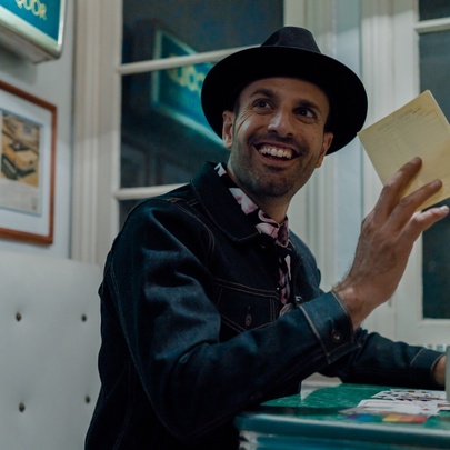 Man wearing a hat, holding a piece of paper above a cup at a table.