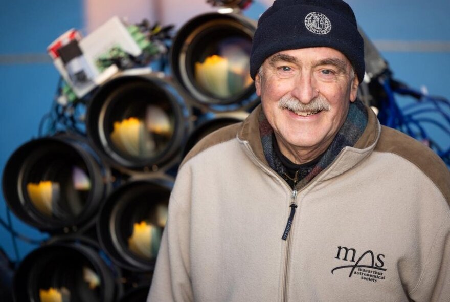 Man wearing a zip-up jacket and beanie standing next to a collection of large-lense telescopes.