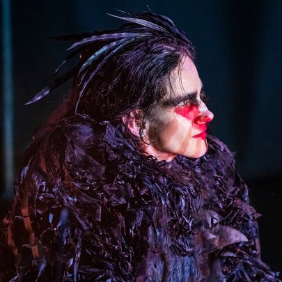 A performer in a dark, textured feathered costume with red face paint on a dark stage.