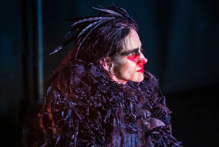 A performer in a dark, textured feathered costume with red face paint on a dark stage.