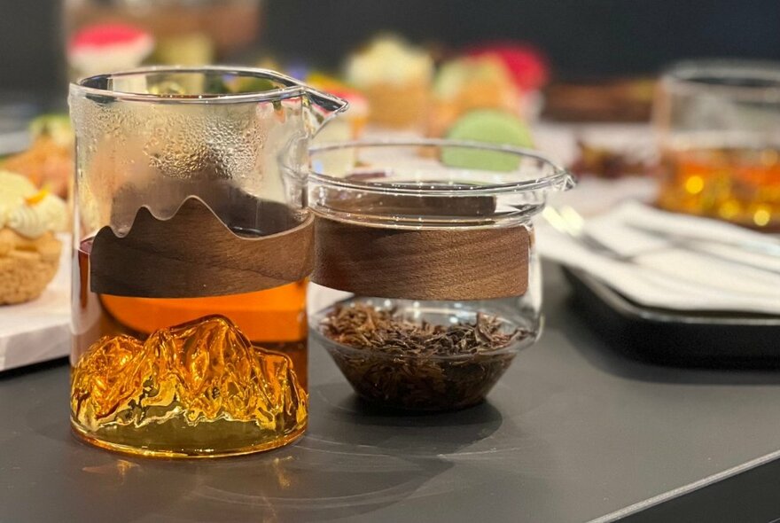 A glass carafe of hot tea and a glass of tea leaves, both with bentwood handles on a black table with treats and other teas in the background. 