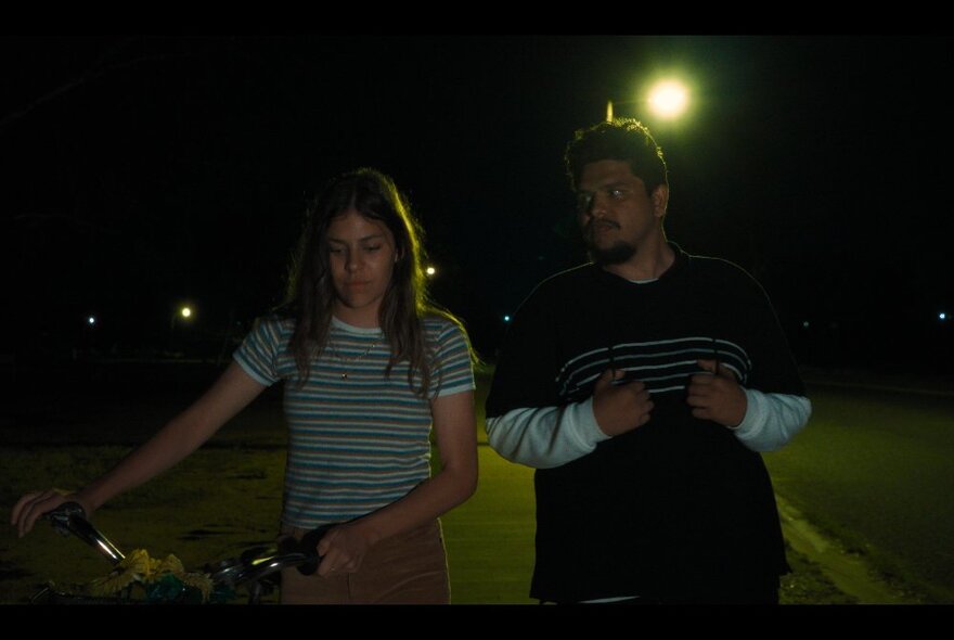 Still from the film Under Streetlights of two actors walking along a street at night, one pushing a bicycle.