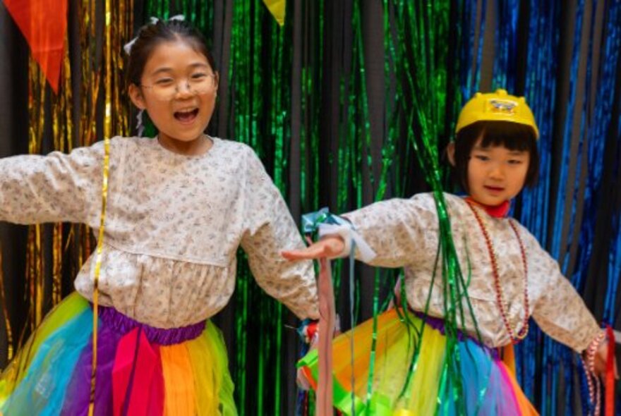 Children wearing rainbow-themed skirts, dancing under tinsel.