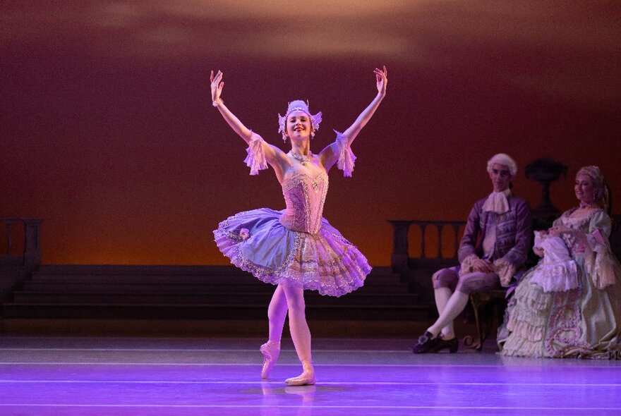 A ballet dancer on a large stage, wearing a tutu and holding a pose with two arms stretched high above her, with two people in costumes seated behind her.