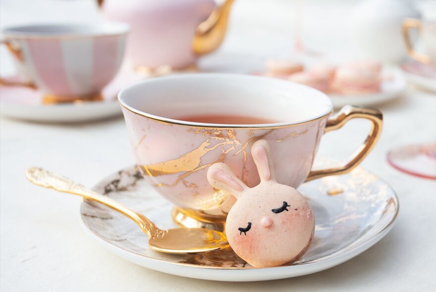 A pink tea cup with a gold handle and a small bunny-shaped treat on a matching saucer, with a gold spoon and a pink teapot in the background.